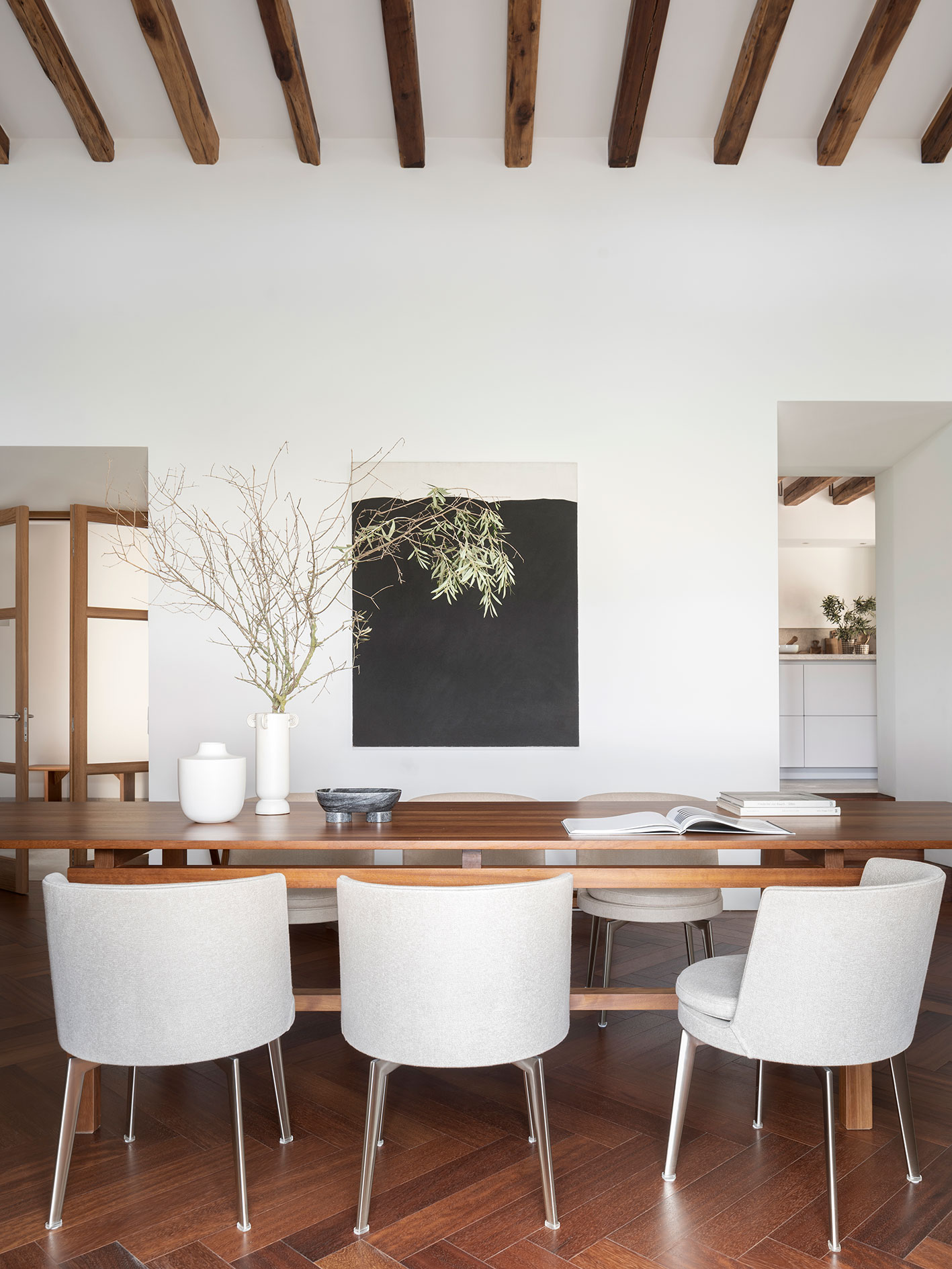 Detalle de la mesa del comedor con sillas grises, cuadro negro, ramas en jarrón y cocina al fondo