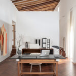 Zona de despacho con escritorio de madera, portátil, lámpara gris, obras de arte y techo de vigas inclinado