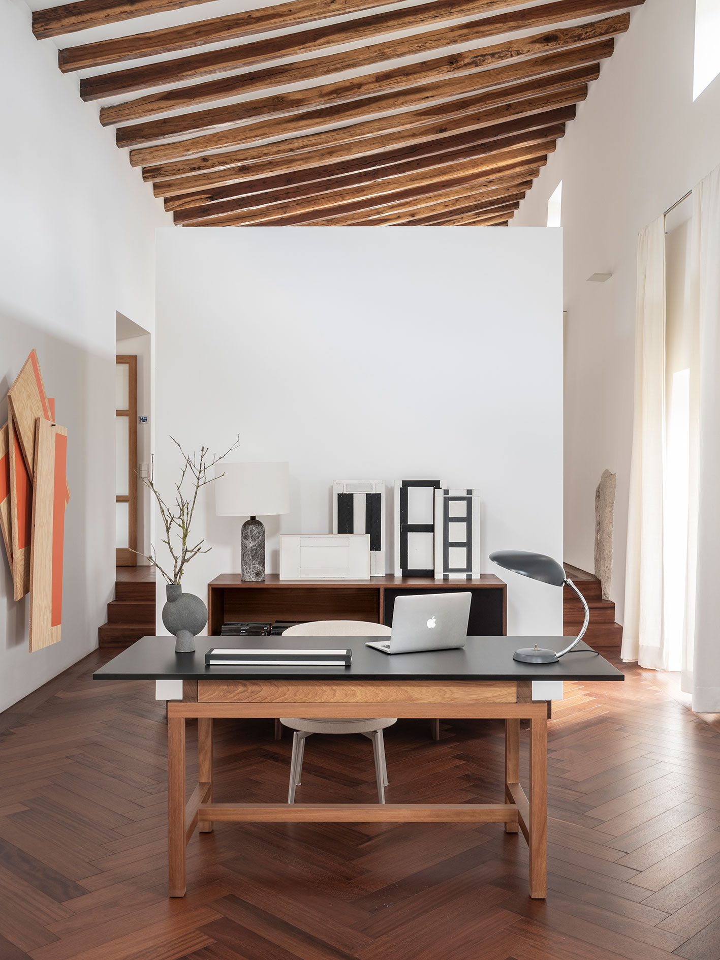 Zona de despacho con escritorio de madera, portátil, lámpara gris, obras de arte y techo de vigas inclinado