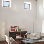 Rincón de trabajo con ventanas pequeñas, aparador de madera con cuadros, sillón claro y lámpara de papel