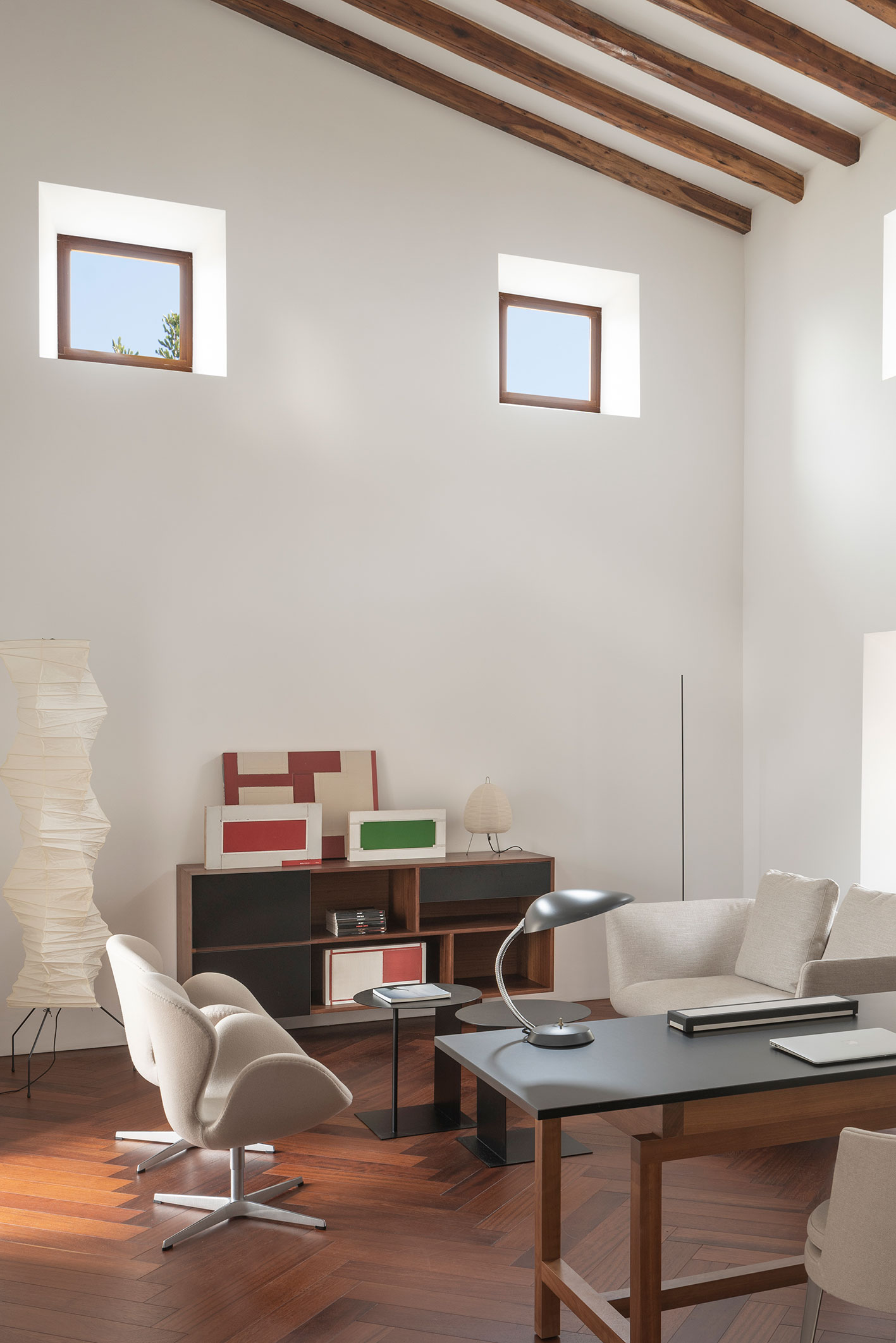 Rincón de trabajo con ventanas pequeñas, aparador de madera con cuadros, sillón claro y lámpara de papel