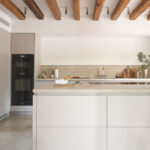 Cocina con isla color crema y encimera de piedra, hornos empotrados, vigas de madera y suelo de piedra