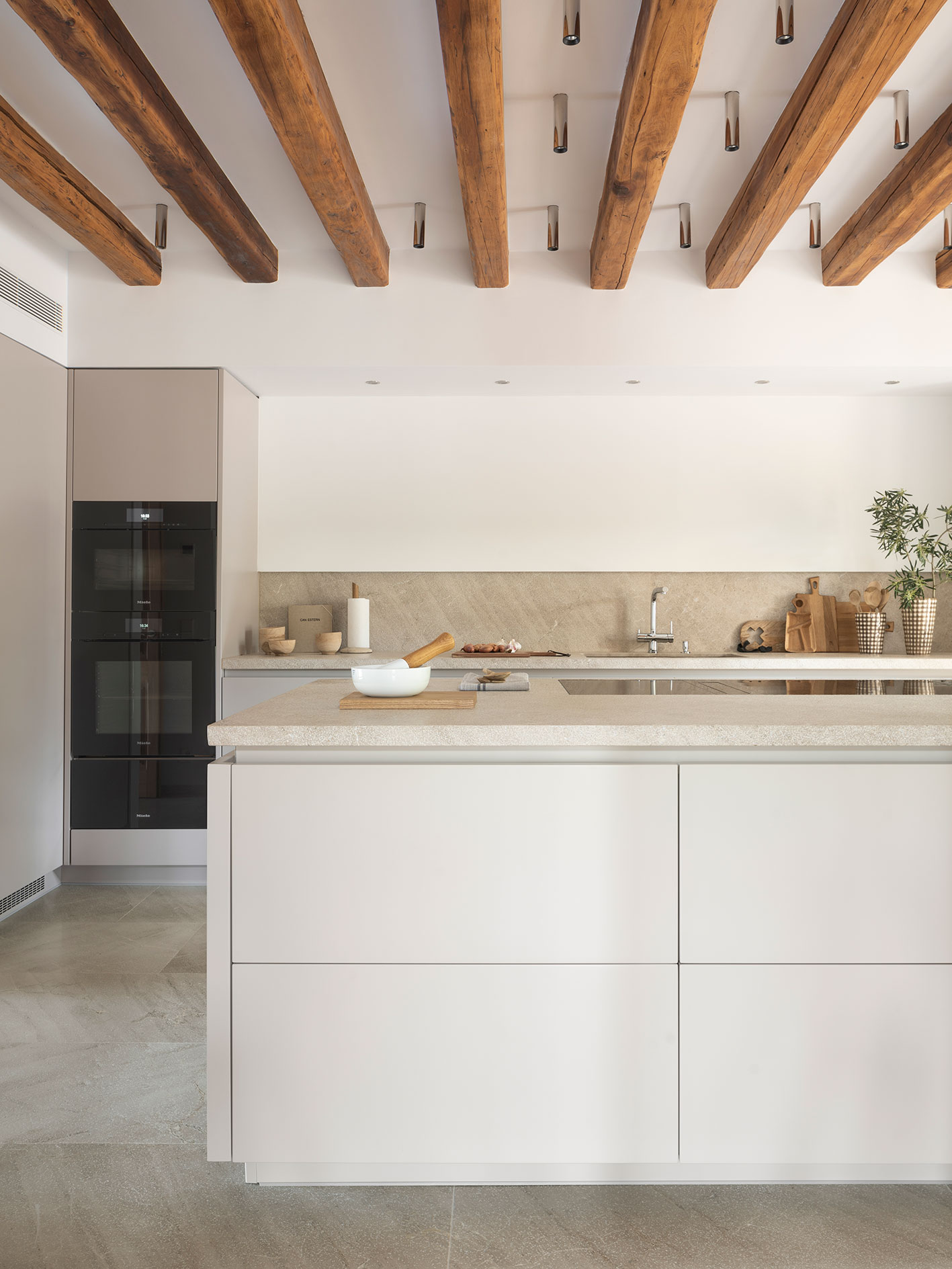 Cocina con isla color crema y encimera de piedra, hornos empotrados, vigas de madera y suelo de piedra