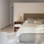 Dormitorio con cama de cabecero tapizado marrón, mesita de madera, lámpara de papel y vigas blancas