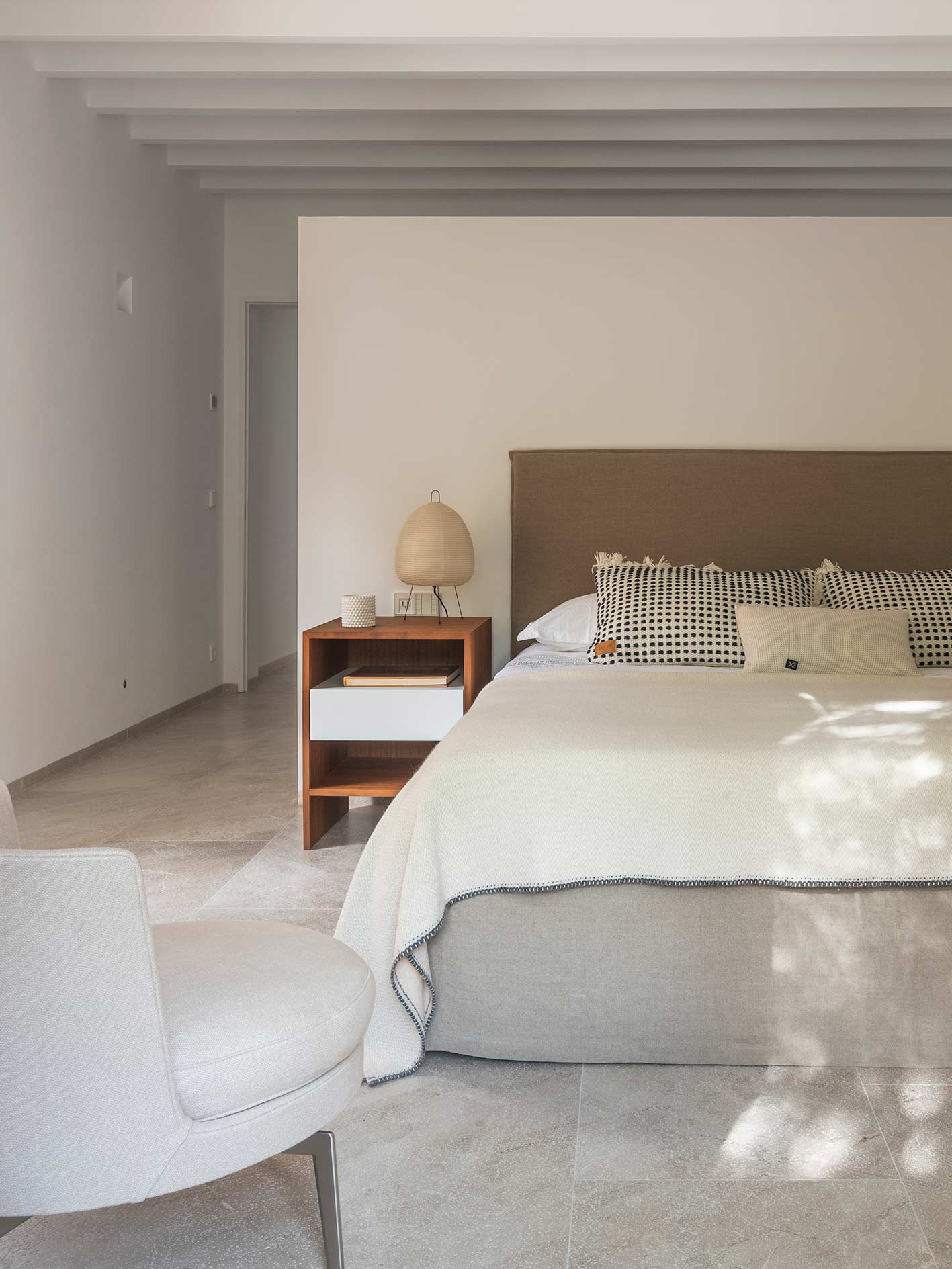 Dormitorio con cama de cabecero tapizado marrón, mesita de madera, lámpara de papel y vigas blancas
