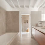 Baño con bañera revestida en piedra beige, doble lavabo, mueble de madera y ducha al fondo
