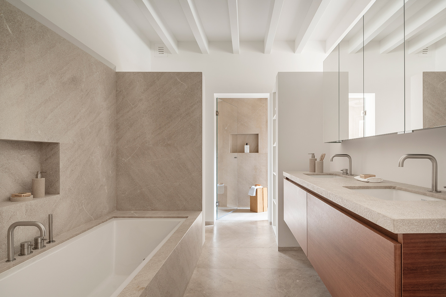 Baño con bañera revestida en piedra beige, doble lavabo, mueble de madera y ducha al fondo