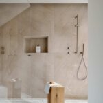 Ducha revestida de piedra beige con vetas, mampara de cristal, teleducha y taburete de madera con toalla