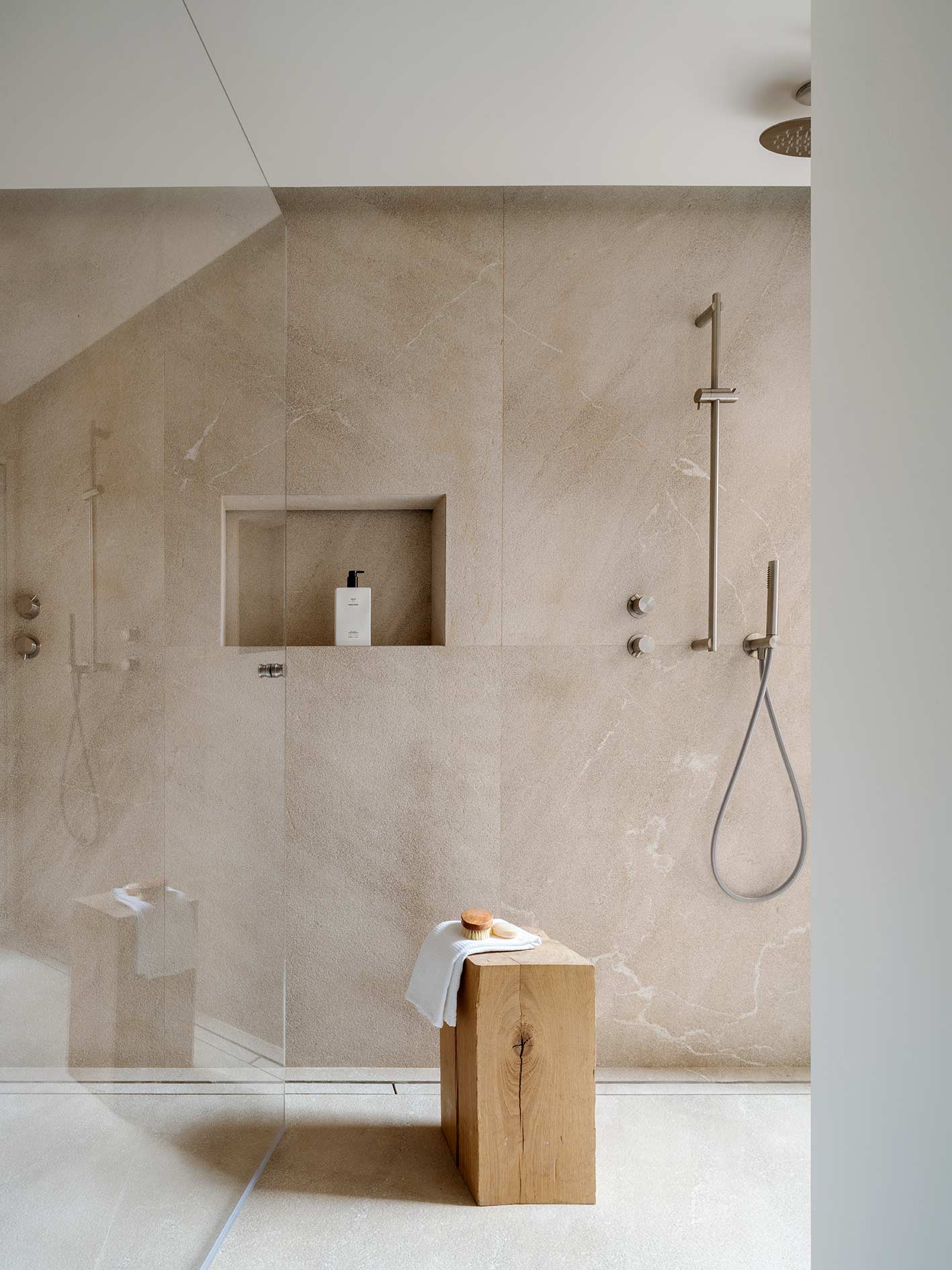 Ducha revestida de piedra beige con vetas, mampara de cristal, teleducha y taburete de madera con toalla