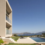 Beige Fassade mit Glasbalkonen neben Pool und Garten, dahinter Meer und Berge