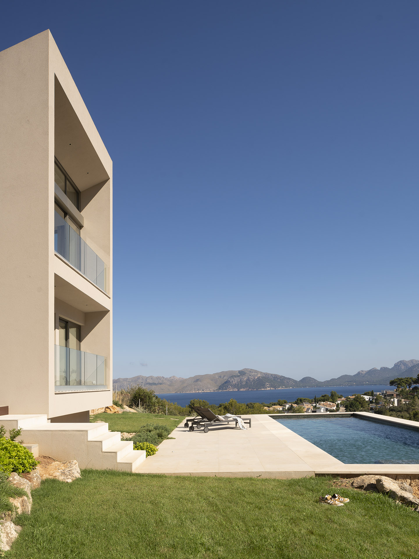 Beige Fassade mit Glasbalkonen neben Pool und Garten, dahinter Meer und Berge