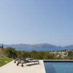 Piscina y tumbonas en terraza de piedra clara con jardín, vistas a la bahía y montañas de día