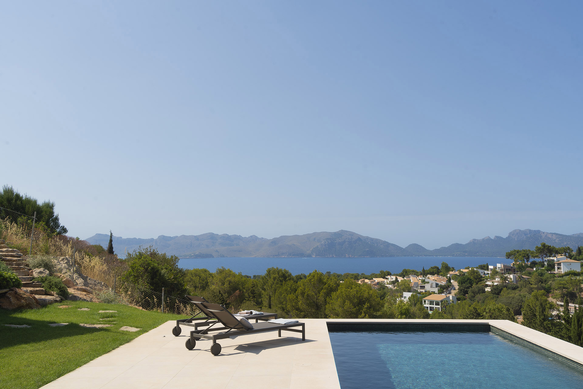 Piscina y tumbonas en terraza de piedra clara con jardín, vistas a la bahía y montañas de día