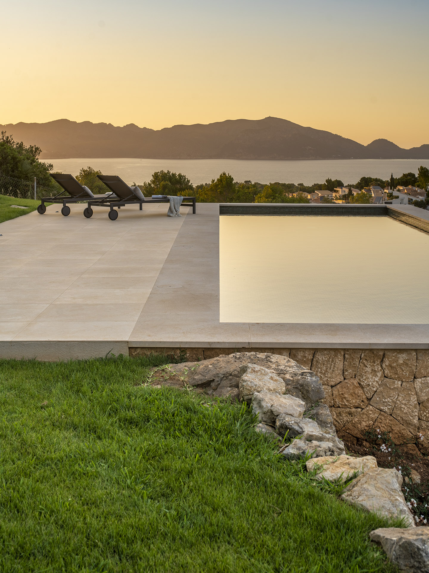 Pool und Sonnenliegen auf hellem Steinbelag bei Sonnenuntergang, dahinter Bucht und Berge