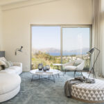 Wohnzimmer mit weißem geschwungenem Sofa, blaugrünem Teppich und großen Fenstern mit Blick auf Meer und Berge