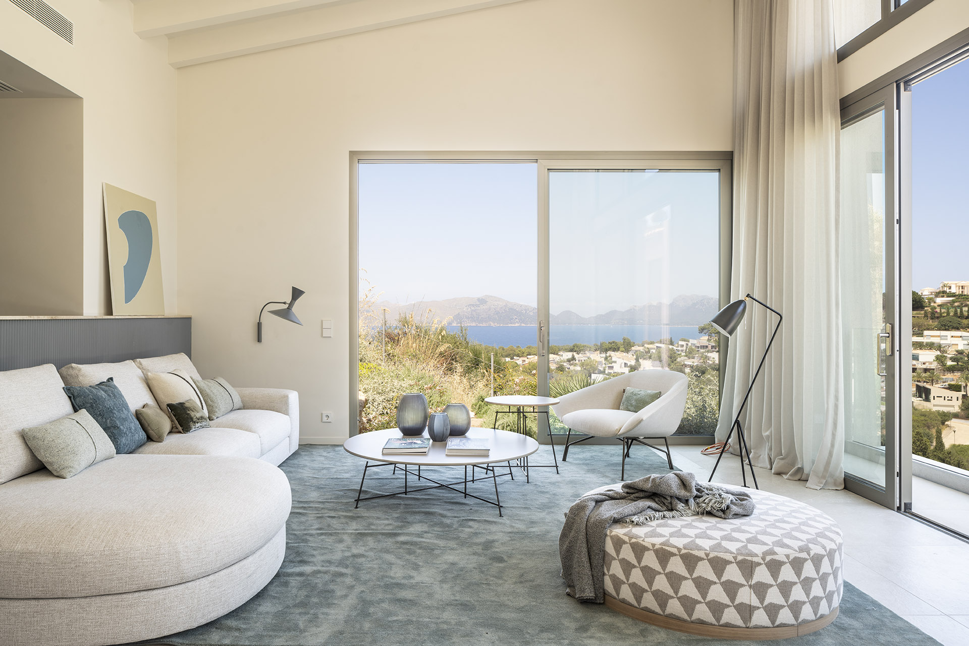 Wohnzimmer mit weißem geschwungenem Sofa, blaugrünem Teppich und großen Fenstern mit Blick auf Meer und Berge