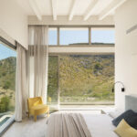 Schlafzimmer mit großem Eckfenster, Voilevorhängen, gelbem Sessel und Blick auf die Berge