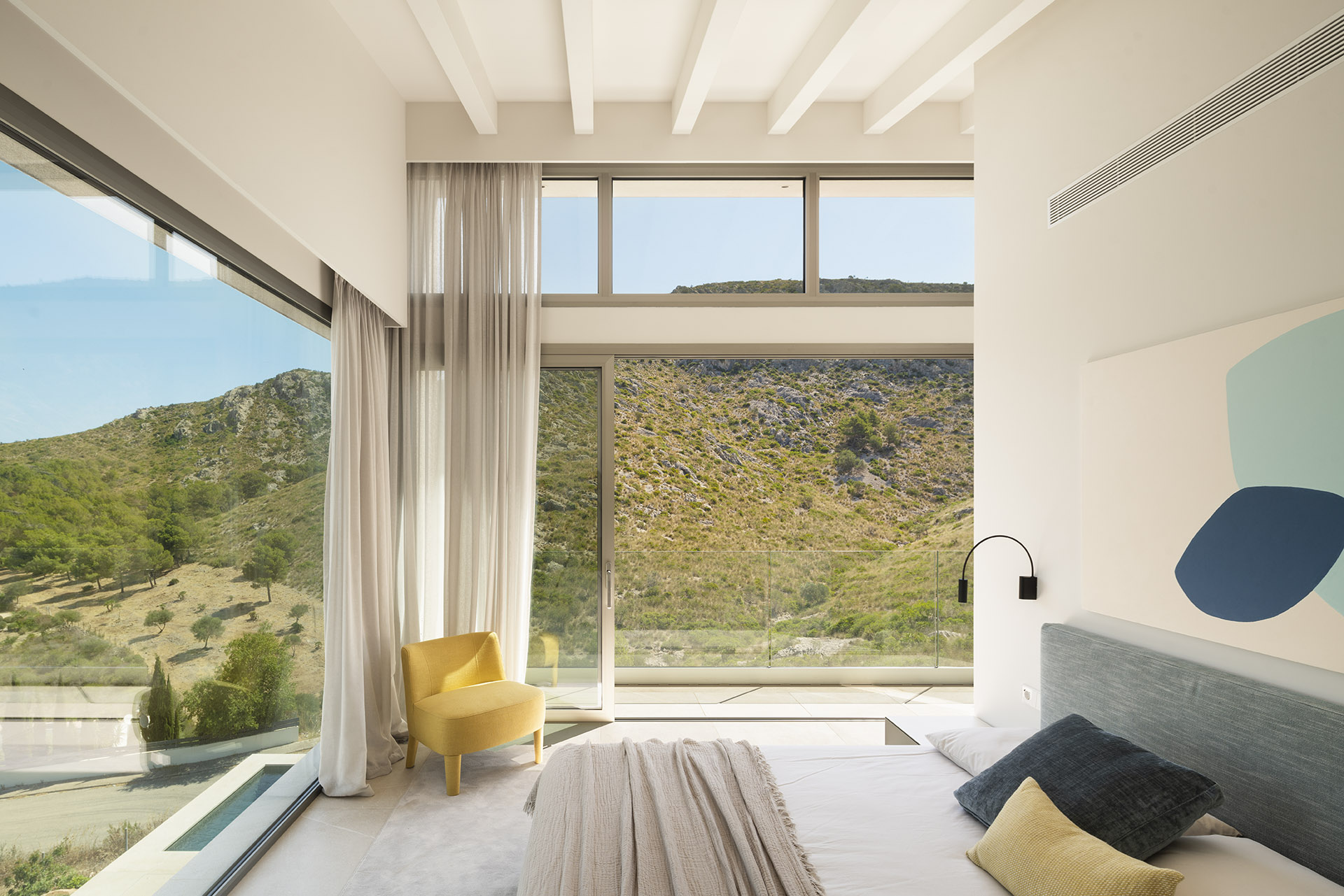 Schlafzimmer mit großem Eckfenster, Voilevorhängen, gelbem Sessel und Blick auf die Berge