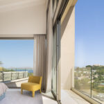 Schlafzimmer mit gelbem Sessel und Vorhängen, zur Terrasse hin offen, mit Blick auf Meer und Wohngebiet