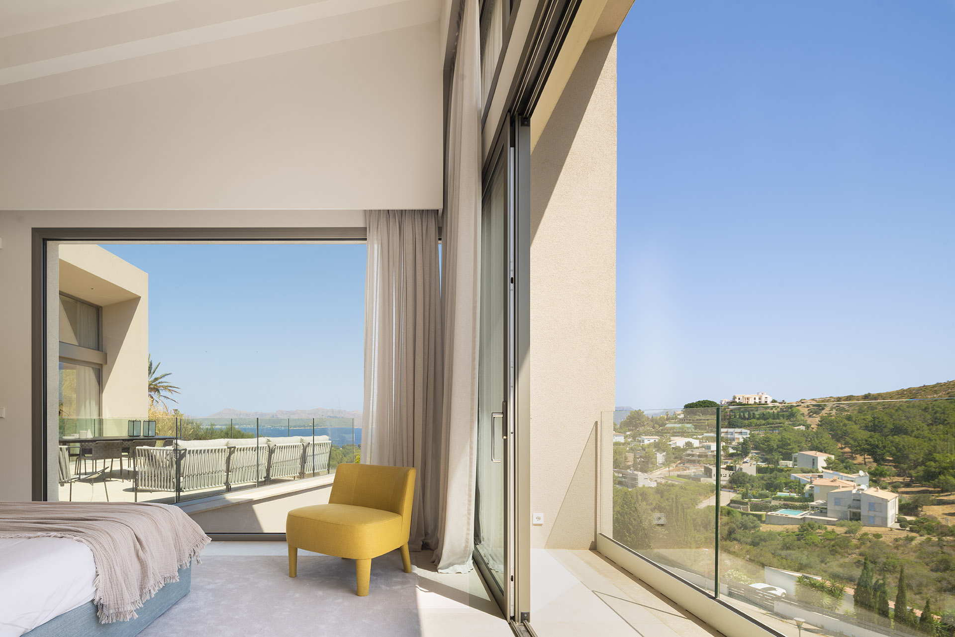 Schlafzimmer mit gelbem Sessel und Vorhängen, zur Terrasse hin offen, mit Blick auf Meer und Wohngebiet