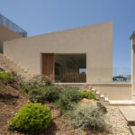 Acceso a la villa con puerta de madera, escaleras entre muros beige y jardín mediterráneo en la ladera