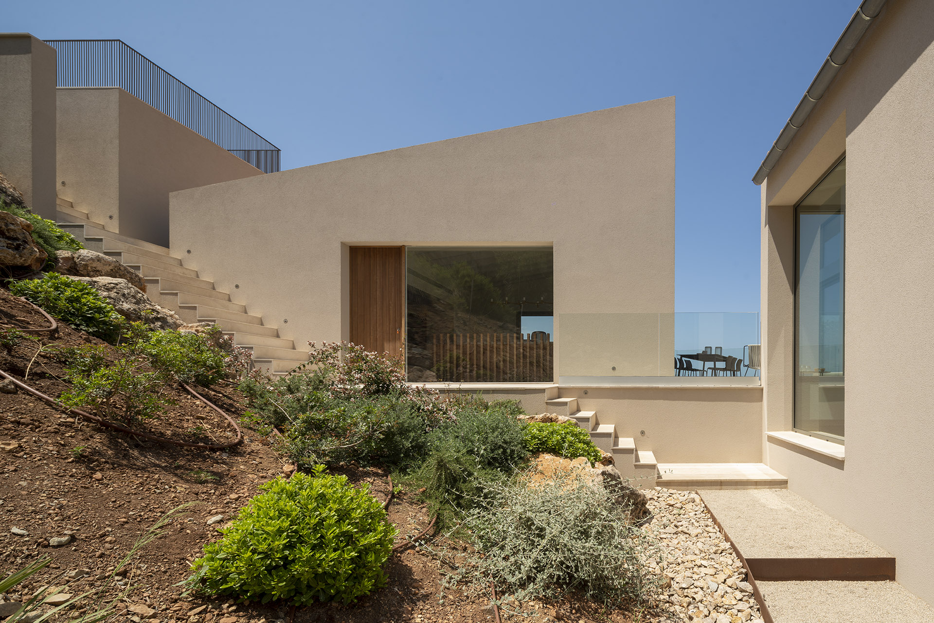 Acceso a la villa con puerta de madera, escaleras entre muros beige y jardín mediterráneo en la ladera