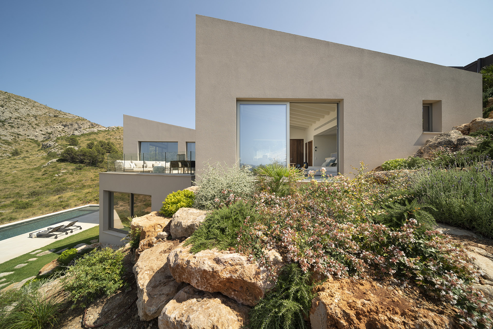 Großes Wohnzimmerfenster zwischen Felsen und blühenden Pflanzen, im Hintergrund der Pool am Hang