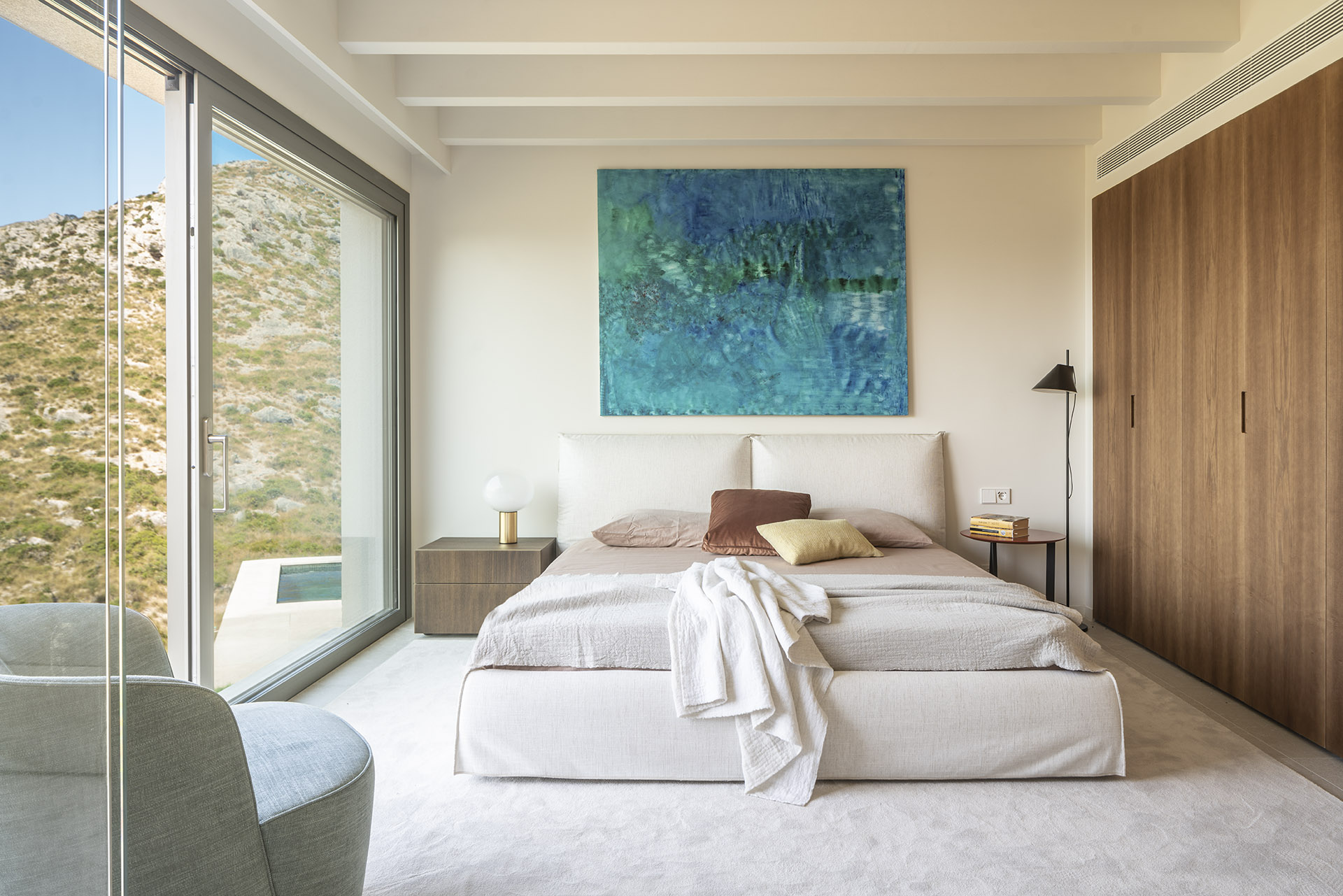 Schlafzimmer mit hellem Kopfteil, großem türkisblauem Gemälde, Holzschrank und großem Fenster zum Hang