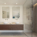 Dusche mit Glastrennwand und grauer Verkleidung neben rechteckigen Spiegeln und Wandarmaturen