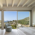 Schlafzimmer mit weißen Balken, großem Fenster mit Blick auf Wohngebiet und Meer sowie hellblauem Sessel