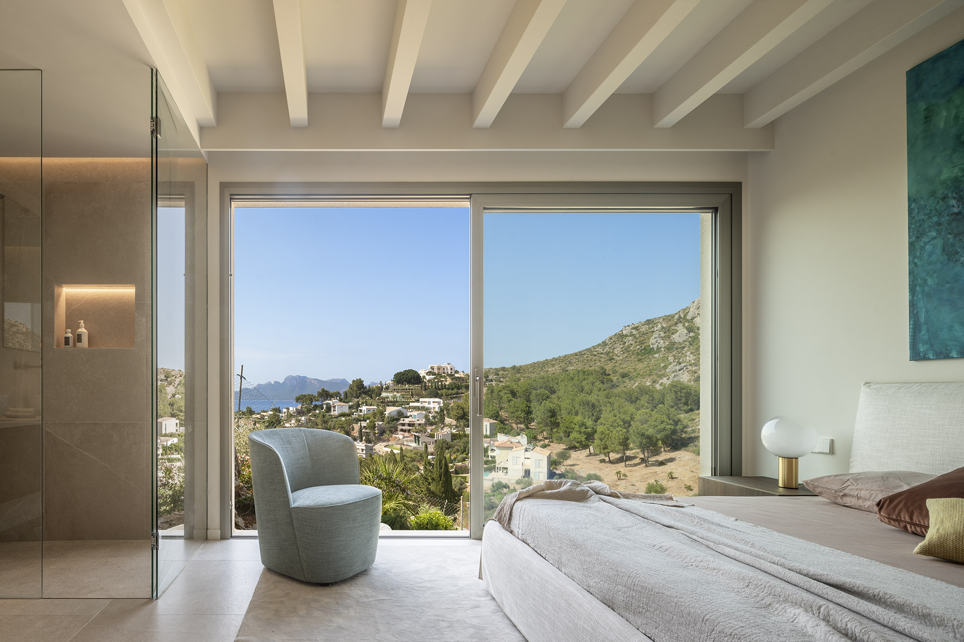 Schlafzimmer mit weißen Balken, großem Fenster mit Blick auf Wohngebiet und Meer sowie hellblauem Sessel