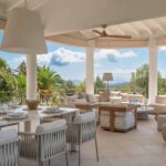 Terraza cubierta con mesa de comedor y sillas tejidas, sofás crema al fondo y palmeras