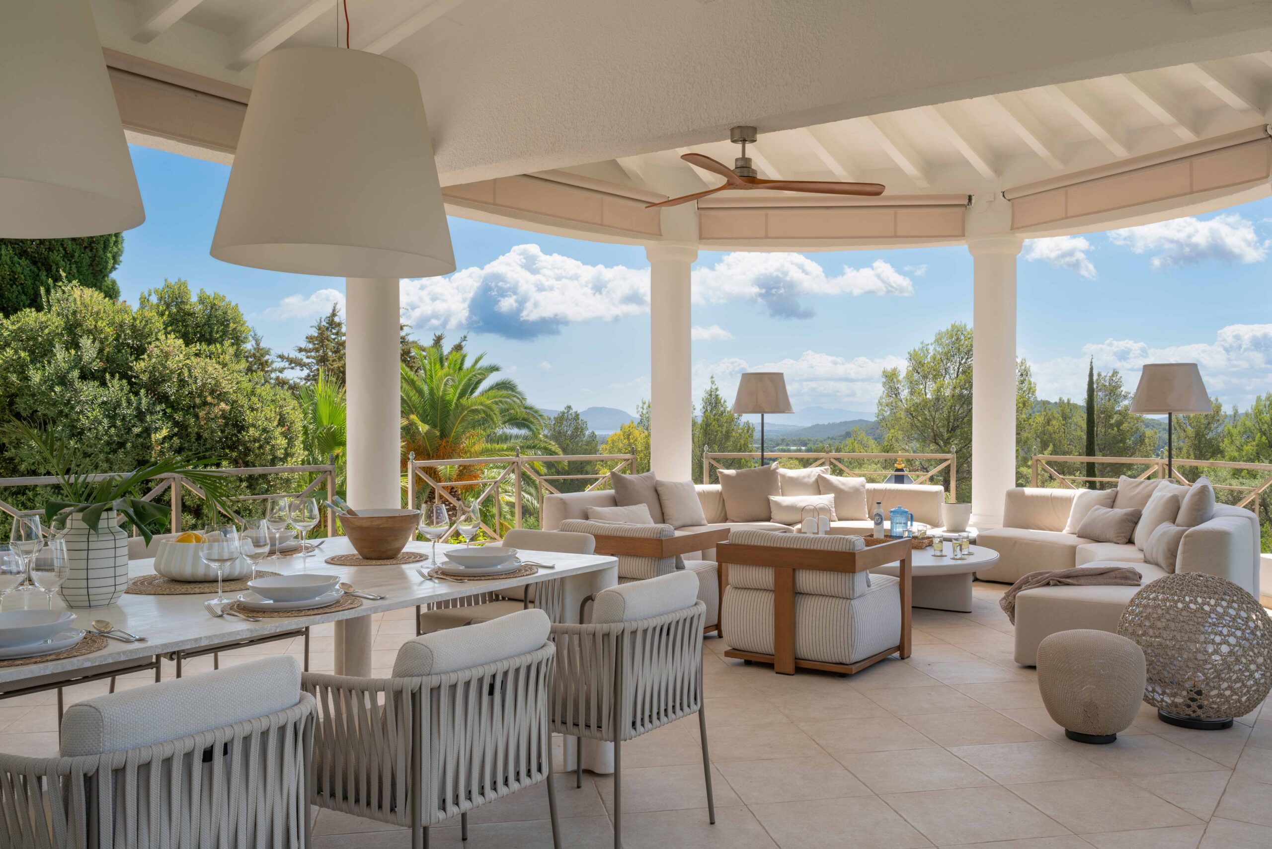 Terraza cubierta con mesa de comedor y sillas tejidas, sofás crema al fondo y palmeras