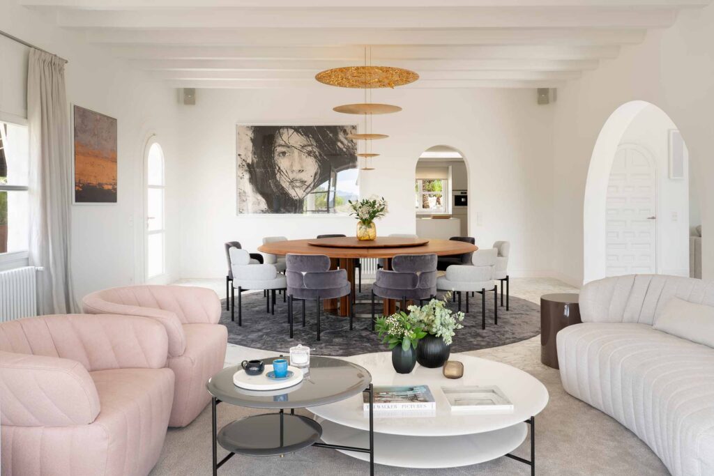 Salón y comedor abiertos con mesa redonda de madera, sillones rosas, mesas de centro y arcos blancos