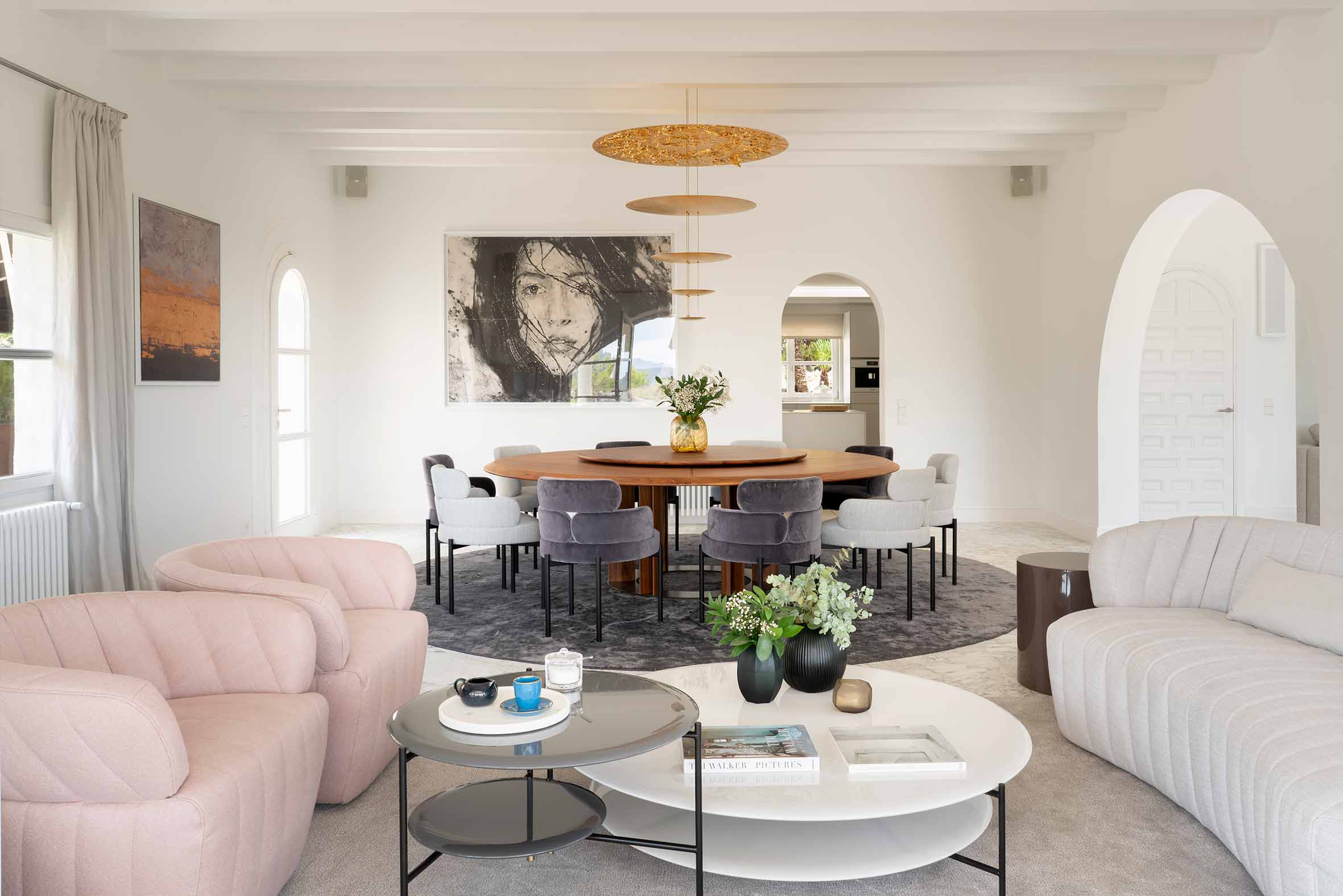 Salón y comedor abiertos con mesa redonda de madera, sillones rosas, mesas de centro y arcos blancos