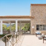 Terraza porticada con fachada de piedra, cubierta de teja, muebles de madera y montañas al fondo