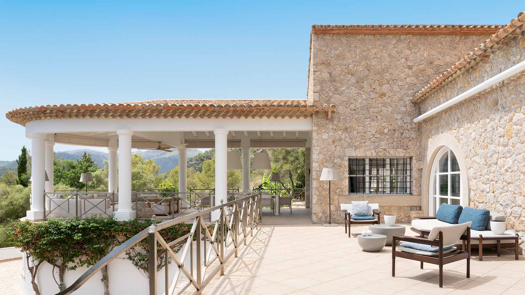Terraza porticada con fachada de piedra, cubierta de teja, muebles de madera y montañas al fondo