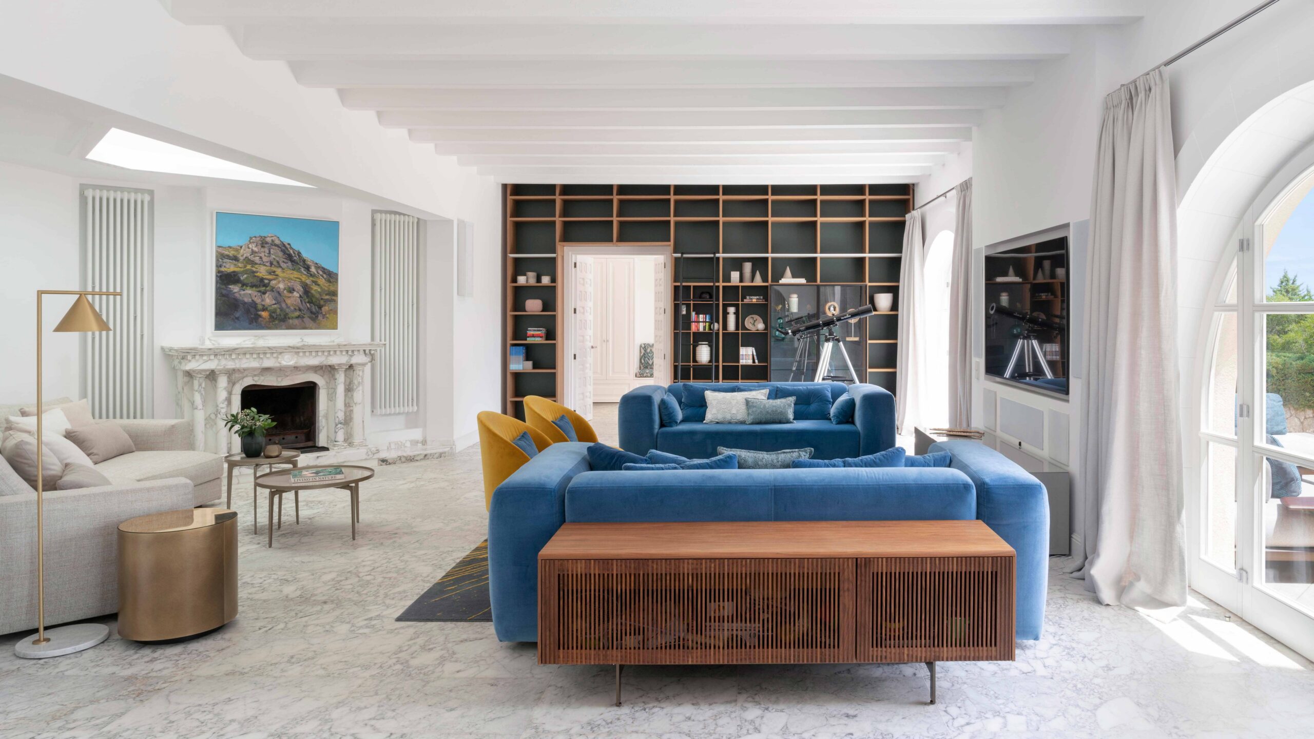 Wohnzimmer mit blauen Samtsofas, Marmorkamin, Bücherregal aus Holz und großem Bogenfenster