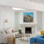 Chimenea de mármol blanco con cuadro de paisaje montañoso, radiadores verticales, sofá beige y mesa lateral dorada