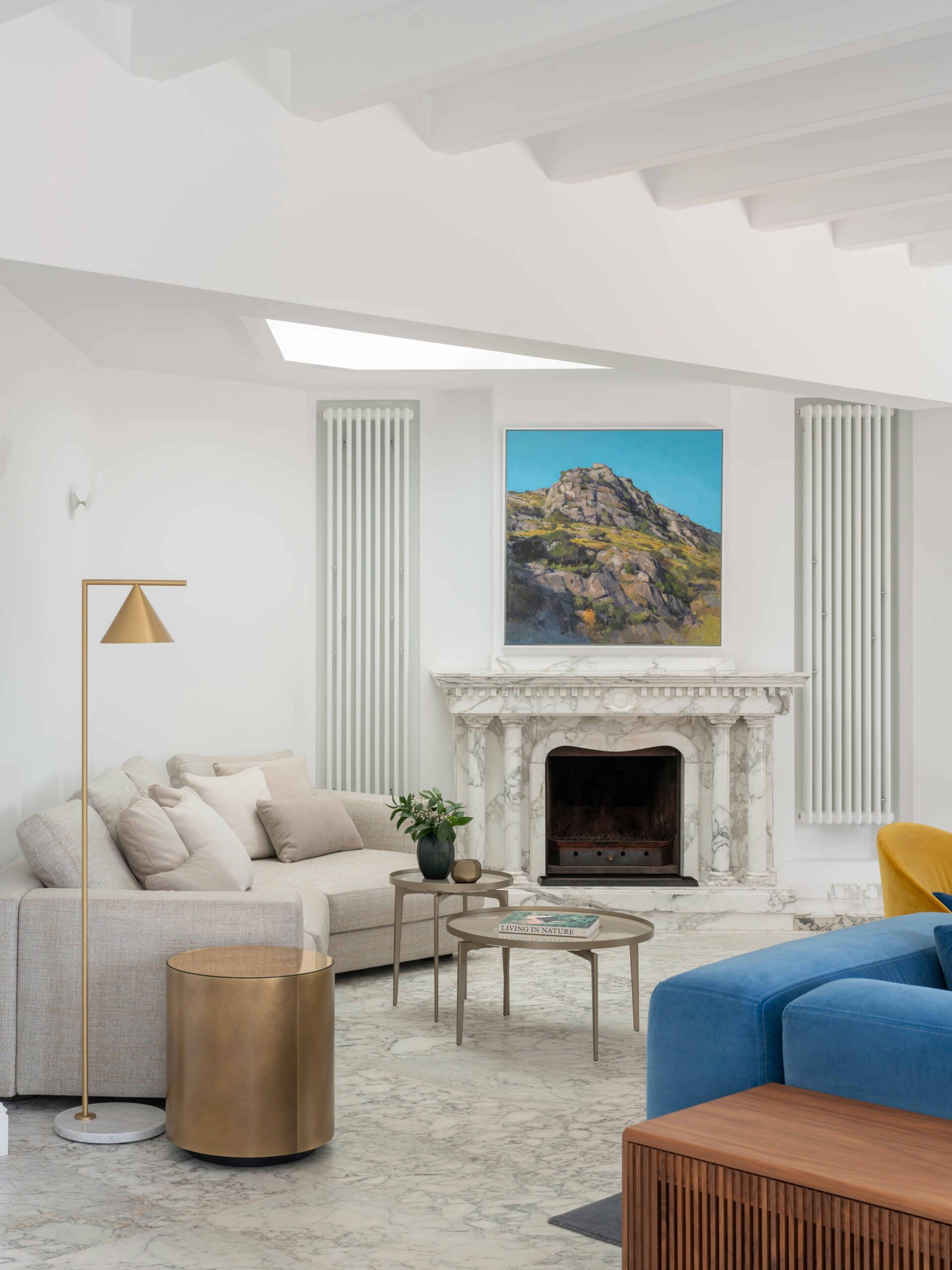 Chimenea de mármol blanco con cuadro de paisaje montañoso, radiadores verticales, sofá beige y mesa lateral dorada
