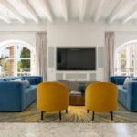 Salón con dos sofás azules, sillones mostaza, alfombra estampada, librería de madera con escalera y telescopio