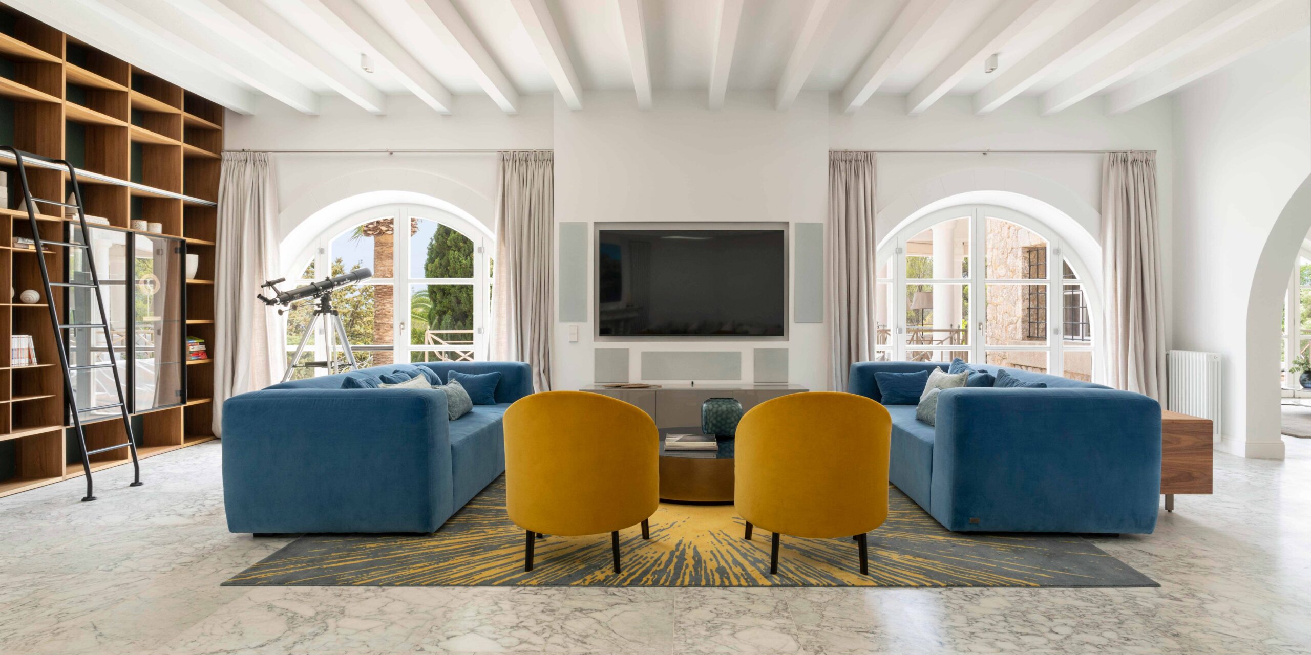 Salón con dos sofás azules, sillones mostaza, alfombra estampada, librería de madera con escalera y telescopio