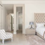 Dormitorio principal con cabecero capitoné, sillón estampado geométrico, armarios blancos y paso al baño