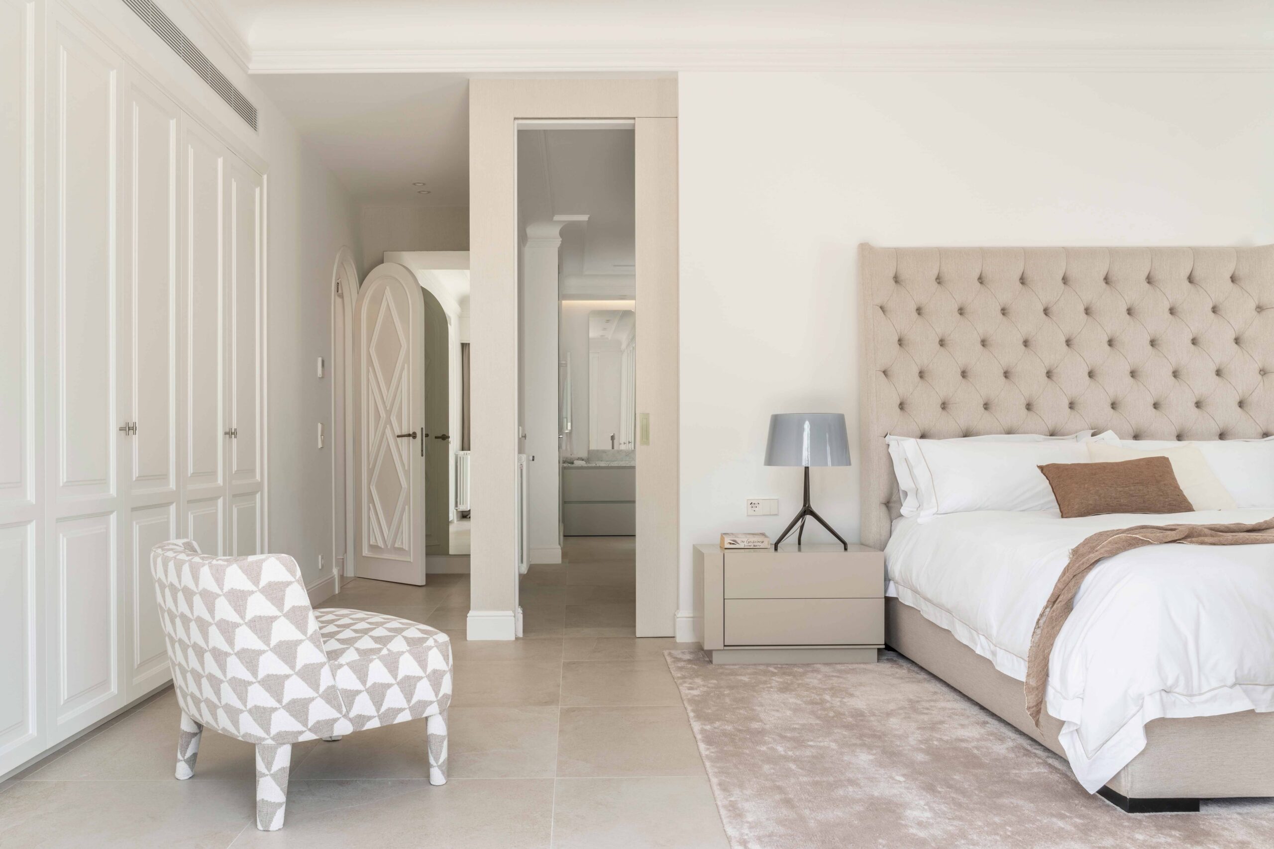 Dormitorio principal con cabecero capitoné, sillón estampado geométrico, armarios blancos y paso al baño