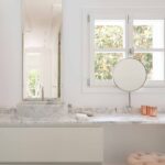 Zona de tocador con encimera y lavabo de mármol, espejo redondo, ventana y puf rosa capitoné