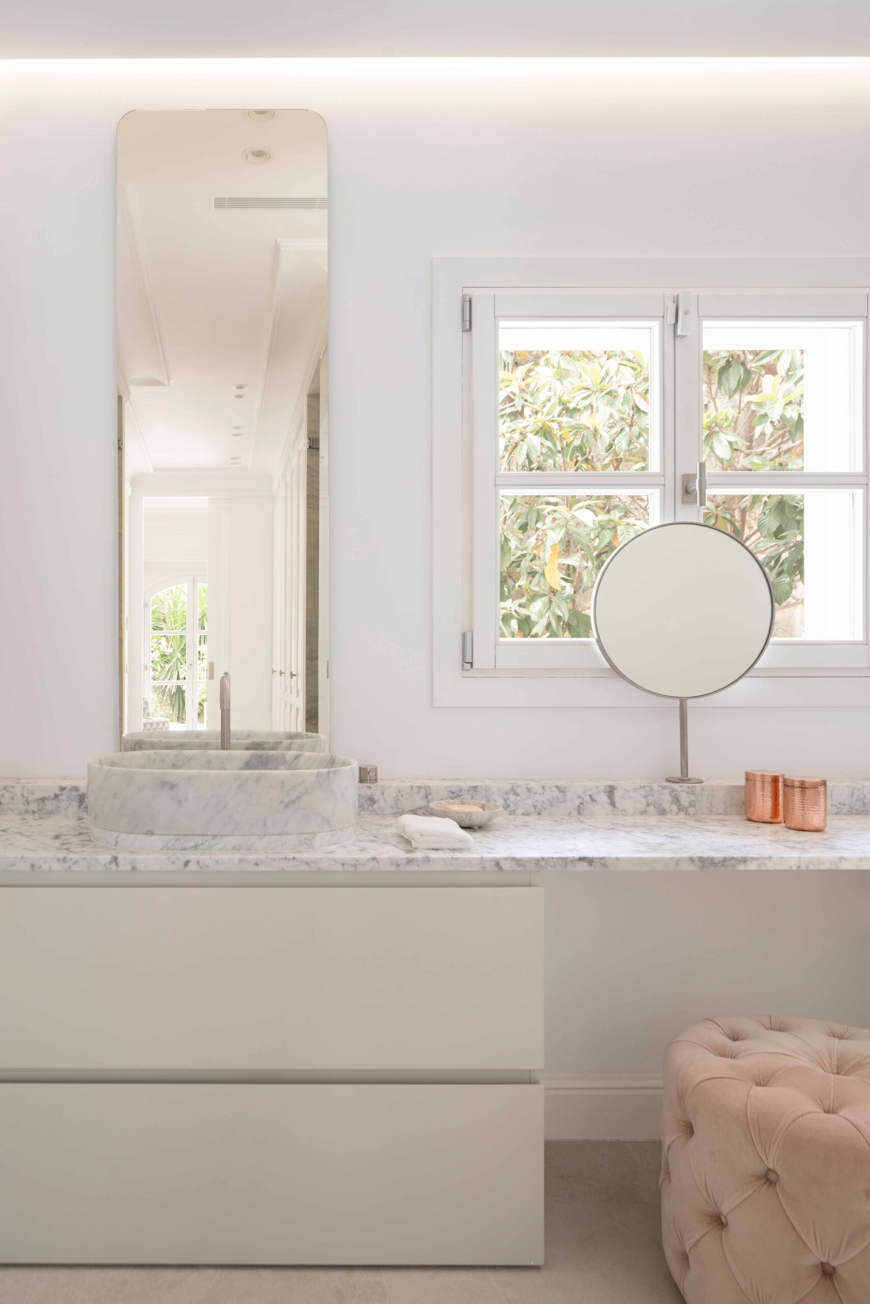 Zona de tocador con encimera y lavabo de mármol, espejo redondo, ventana y puf rosa capitoné