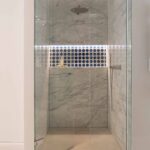 Ducha de mármol con mampara de cristal, hornacina iluminada de azulejos azules y grifería metálica