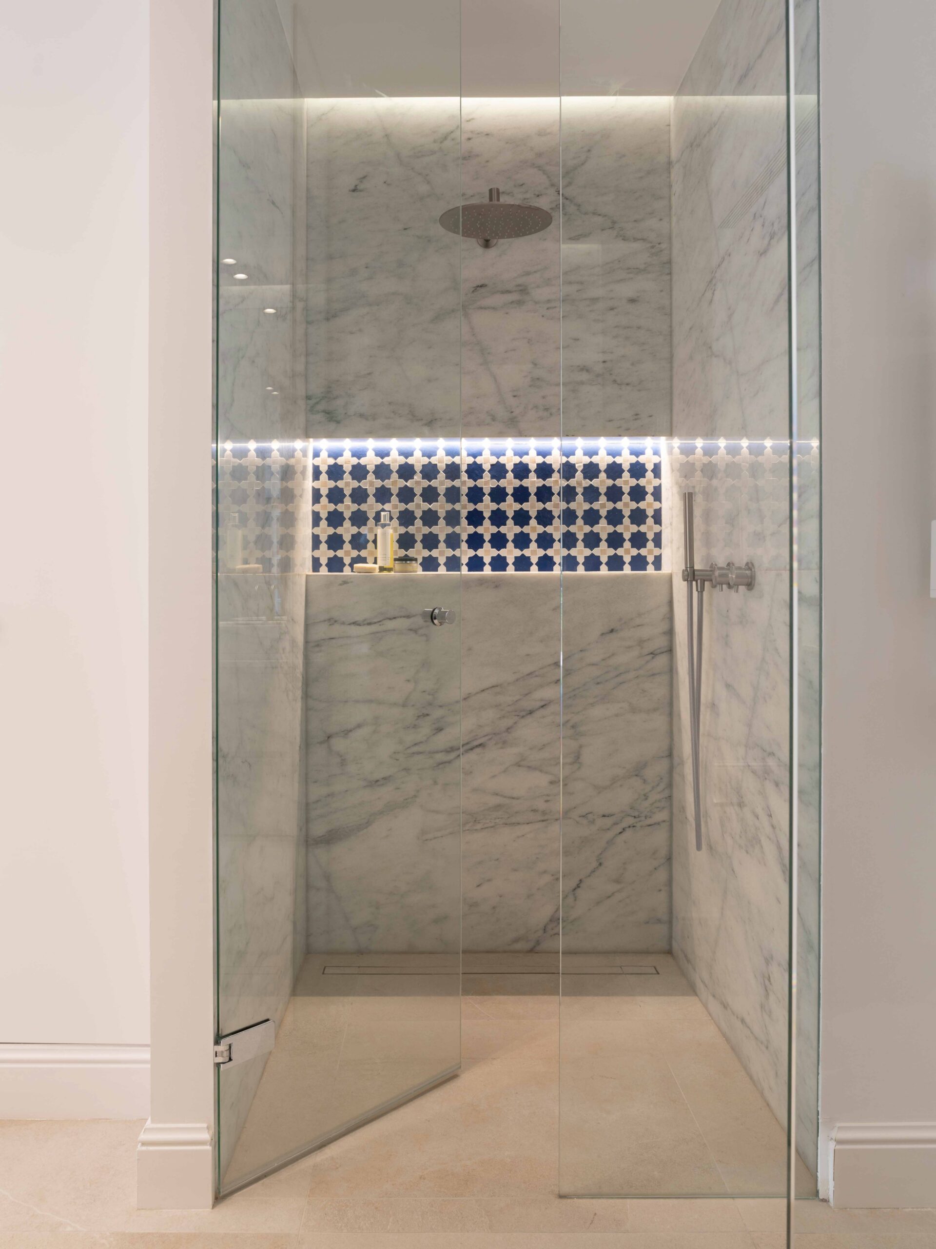 Ducha de mármol con mampara de cristal, hornacina iluminada de azulejos azules y grifería metálica