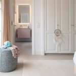 Dormitorio con armarios blancos empanelados, puerta acristalada, puf de tela y baño al fondo