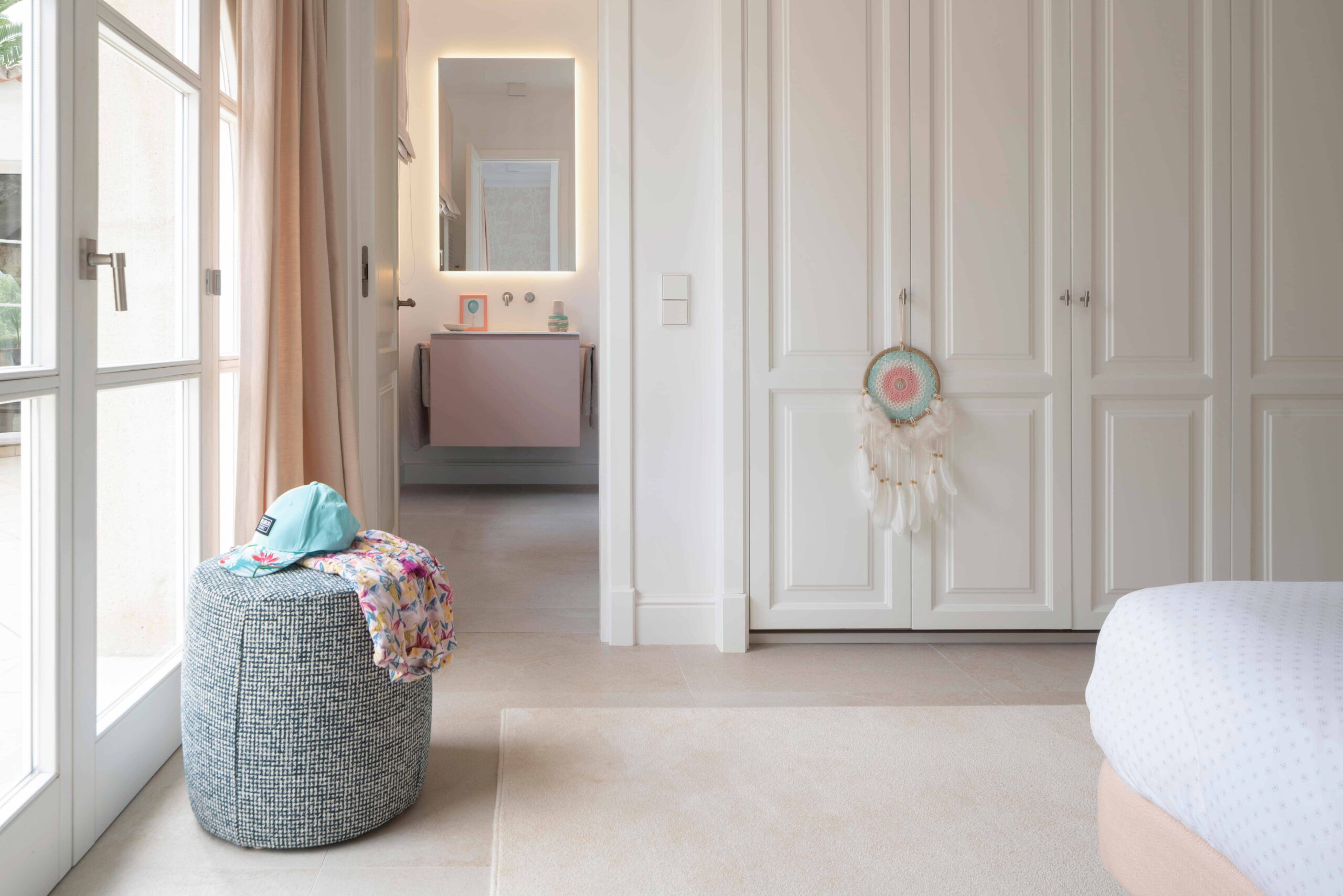 Dormitorio con armarios blancos empanelados, puerta acristalada, puf de tela y baño al fondo